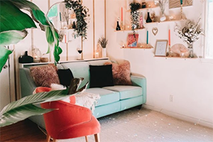 Cozy pastel living room featuring a mint sofa, red accent chair, hanging plants, and sunlit shelves.