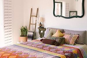 Cozy bedroom with a colorful quilt, throw pillows, ladder shelf, plant, ornate mirror, and sunlit blinds.