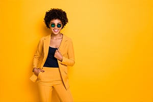 Portrait of a woman in a yellow suit posing against a yellow backdrop, wearing sunglasses and smiling.