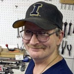 Man in a blue shirt wearing a cap with an L logo, standing in a workshop with tools on a pegboard behind him.
