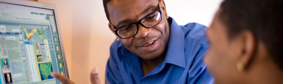 Man wearing glasses in a blue shirt explains data displayed on a computer screen to a colleague.