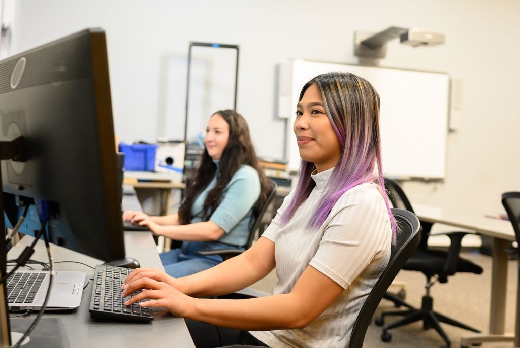 Smiling woman with purple-dyed hair types at a desktop computer in a bright office, with a colleague in the background.