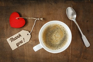 Cup of coffee on a wooden table with a red heart and a 'Thank you!' tag beside a spoon.