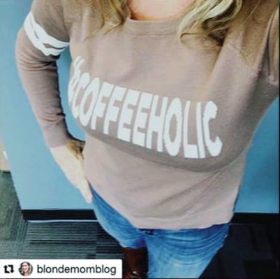 Woman wearing a beige long-sleeve shirt that reads COFFEEHOLIC, standing with one hand on her hip.