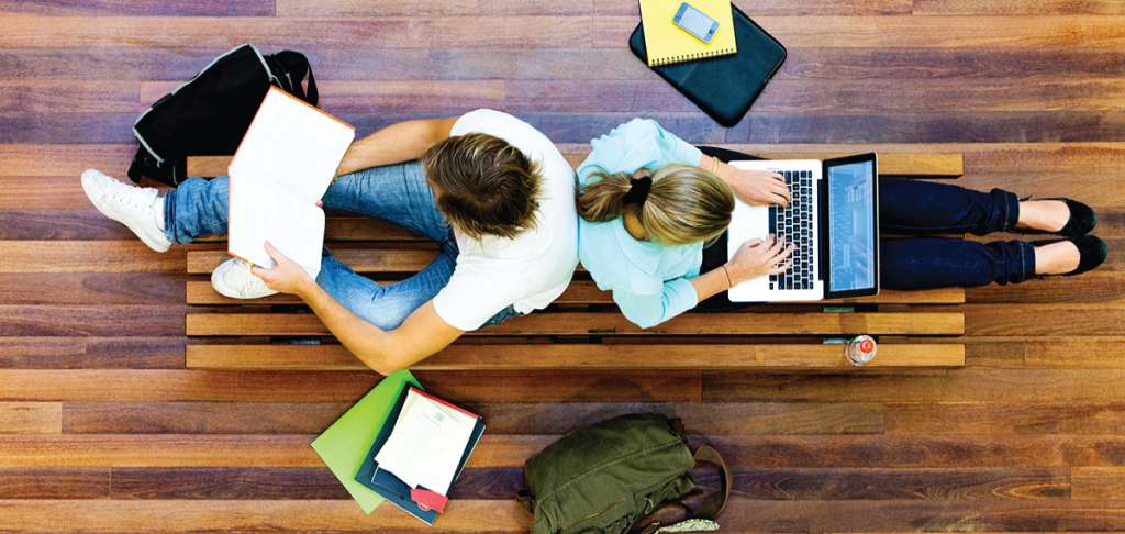 Overhead view of two students studying on a wooden bench: one with a laptop, the other with a book.
