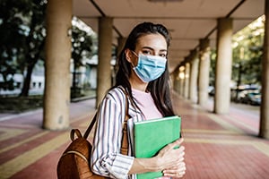 Student wearing a blue face mask walks along a covered campus walkway, carrying a green notebook and a backpack.