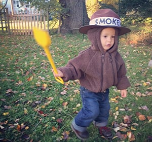 Toddler in a brown hooded jacket holding a yellow shovel in a leaf-covered yard.
