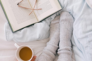Cozy bed scene with an open book, gray knit socks, a mug of tea, and wooden knitting needles.