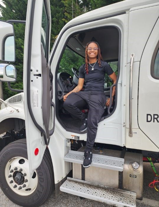 Smiling person sits on the metal steps of an open white truck cab, wearing a black outfit.