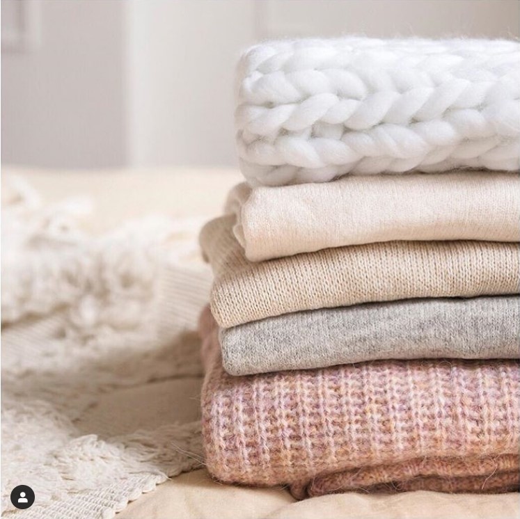 Stack of folded knit sweaters in cream, beige, gray, and pink tones.
