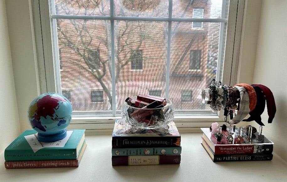 Globe on a stack of books on a bright windowsill, glass bowl center, bracelets on the right, with a brick building visible outside.
