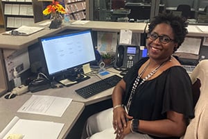 Smiling woman sits at a cluttered office desk with a computer, phone, and papers.