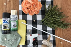 Craft supplies arranged on a black-and-white checkered cloth: orange roses, faux greenery, glue gun, scissors, tape, and spray paint.