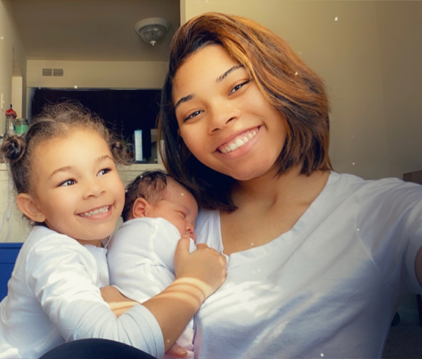 Young woman smiles for a selfie with her daughter holding a newborn baby in a bright home.