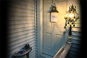 Cozy porch entry with weathered siding, a hanging lantern near a white front door, and a small table at the railing.