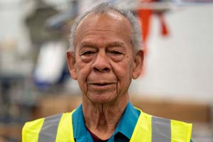 Elderly man wearing a bright yellow safety vest over a blue shirt in a workshop