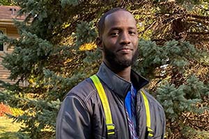 Man wearing a dark jacket with yellow reflective stripes stands outdoors among trees.