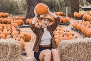 Smiling girl sits among pumpkins at the pumpkin patch, holding a pumpkin above her head.