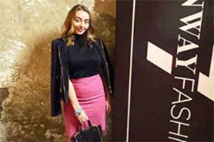 Woman poses in front of a fashion banner wearing a black top, pink pencil skirt, and a dark blazer.