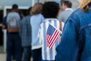 Close-up of a person in a denim jacket holding a small American flag while a line of people stands in the background.