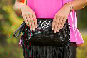 Hands holding a black fringed snakeskin handbag with a triangular emblem, paired with a bright pink outfit.