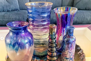 Decorative collection of iridescent glass vases and candleholders in blue, purple, and pink tones on a reflective surface.