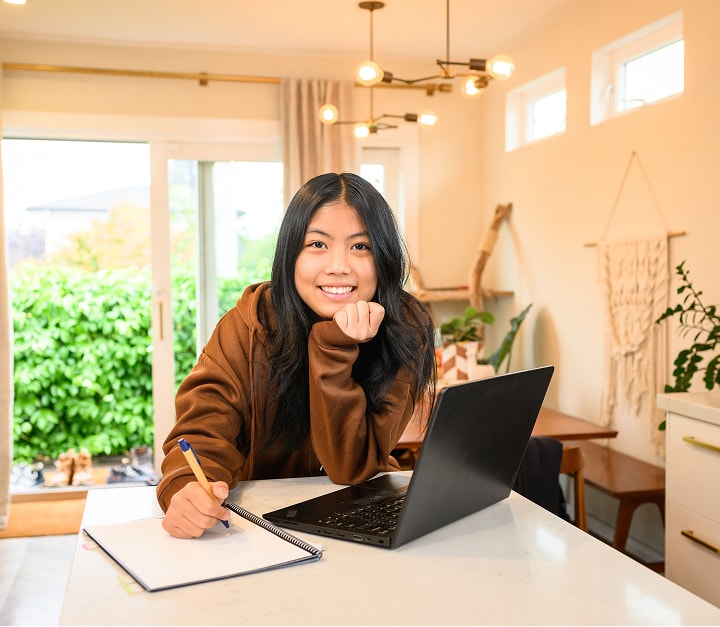 Young woman sits at a bright kitchen island, smiling while writing in a notebook with a laptop nearby.