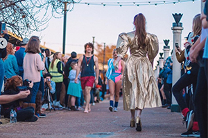 Model walking down an outdoor street runway while spectators line both sides.