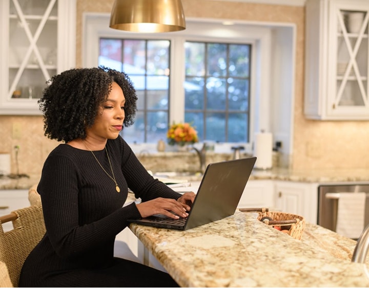 Woman with curly hair wearing a black sweater typing on a laptop at a granite kitchen island.
