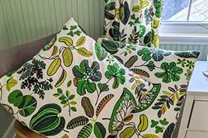 Botanical leaf print cushions and a matching table runner on a white surface in a sunlit, pastel-green corner.