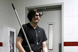 Person with short dark hair and glasses holds a long silver pole in a hallway.