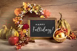 Autumn still life with pumpkins, apples, gourds, a decorative wreath, and a chalkboard reading Happy Fallidays.