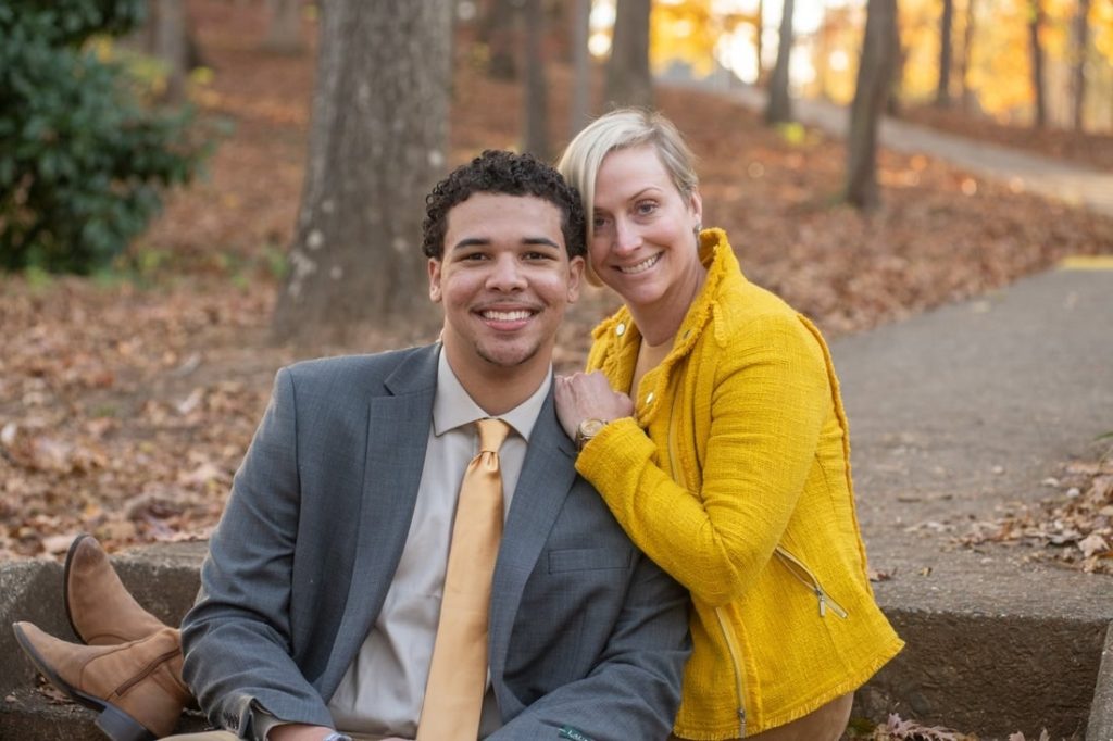 Smiling couple relaxing on a park wall in autumn; man in a gray suit with a yellow tie, woman in a bright yellow jacket.