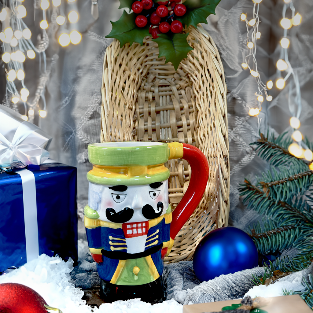 Colorful nutcracker mug in a holiday still life with a wicker vase, blue gift box, blue ornament, pine branches, and twinkling fairy lights.