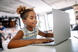 Young girl with curly hair in denim overalls typing on a laptop at a bright classroom table.