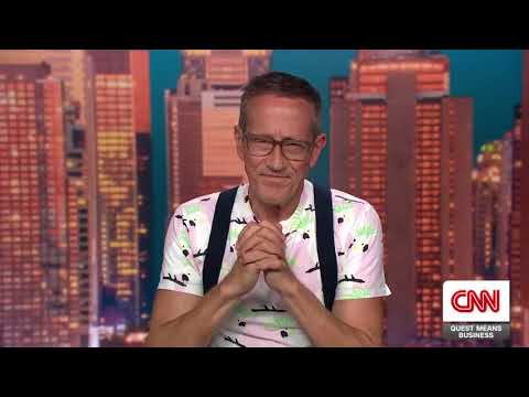 Man with glasses and suspenders sits in a TV studio, hands clasped, city skyline backdrop.