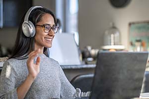 Woman wearing headphones and glasses smiles while using a laptop in a home office.