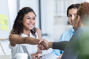 Smiling woman in a white blouse shakes hands with a colleague in a bright office meeting.