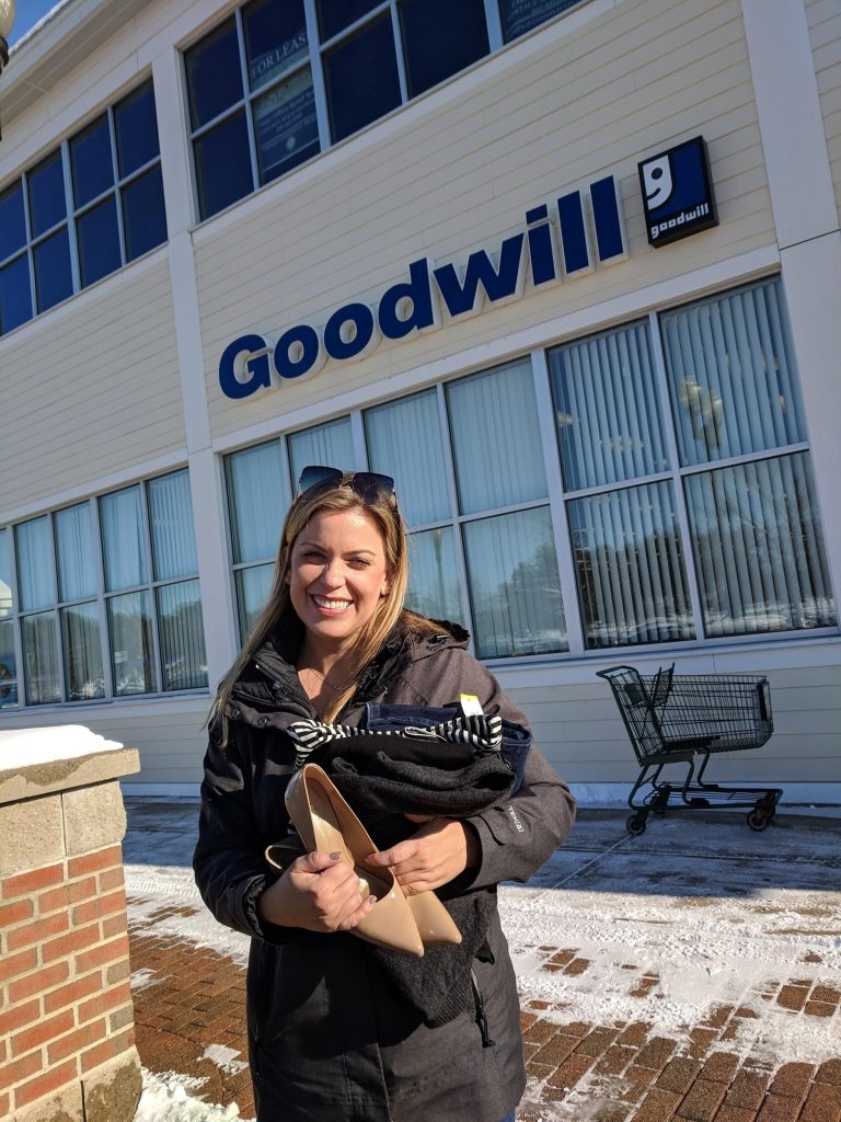 Smiling shopper outside a Goodwill store, holding beige high-heel shoes and bundled clothing.