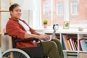 Person in a wheelchair sits at a bright home office desk with a laptop, books, and plants nearby.