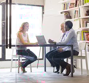 Three professionals sit around a table in a modern, bright office; a woman on the left uses a laptop while two men converse.