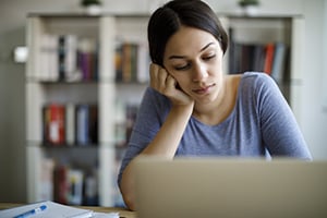 Woman seated at a desk, resting her chin on her hand while using a laptop.