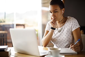 Woman in a white polka-dot shirt sits at a table with a laptop, thinking, in a bright cafe.