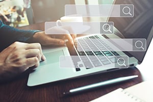 Hands typing on a laptop with floating search bars and magnifying glass icons hovering above the keyboard.