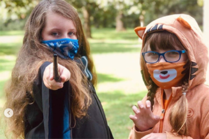 Two children in a park wearing costumes and masks, one pointing a wand toward the camera.