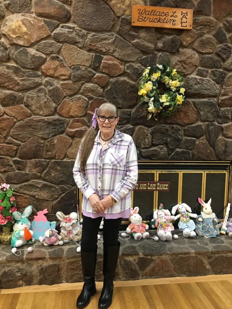 Older woman with glasses and long gray hair in a purple scarf, standing before a stone fireplace with Easter decorations.