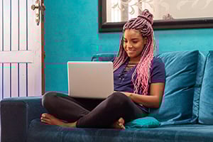 Young woman with pink braided hair sits on a blue sofa, using a laptop in a colorful living room.