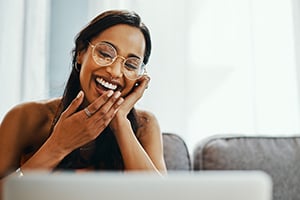 Smiling woman wearing glasses leaning toward a laptop, hands near her mouth in delight.