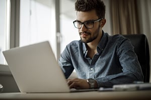 Man with glasses typing on a laptop at a desk in a bright office.