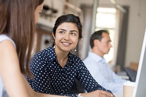 A smiling woman in a navy polka-dot blouse talks with a colleague in an office setting.